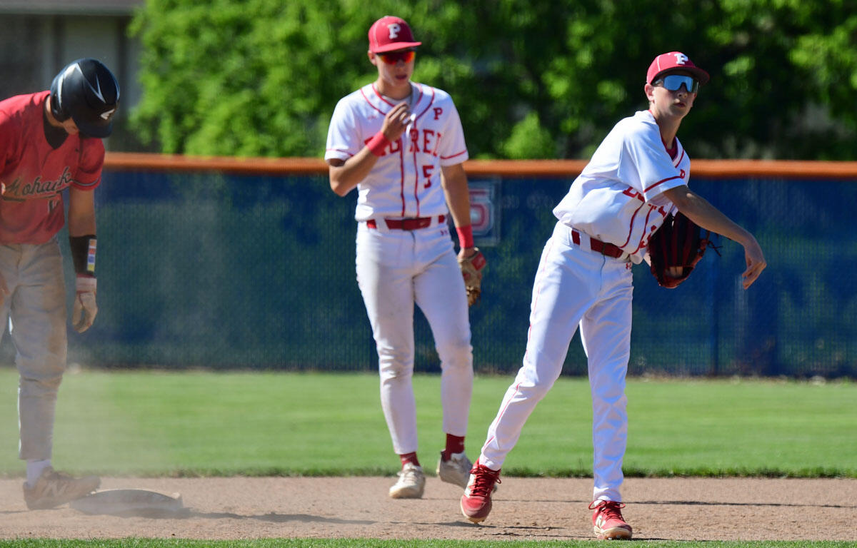 Baseball players on a field: one in a red uniform sliding near a base while two others in white uniforms watch.