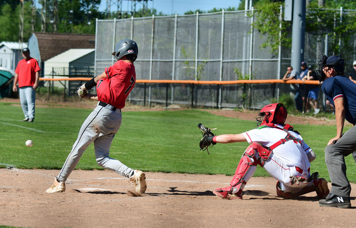 Baseball batter in red swings at a pitch as the catcher in white gear reaches toward the ball at home plate.