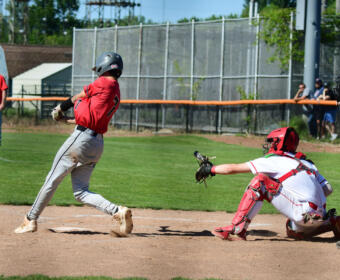 Baseball batter in red swings at a pitch as the catcher in white gear reaches toward the ball at home plate.