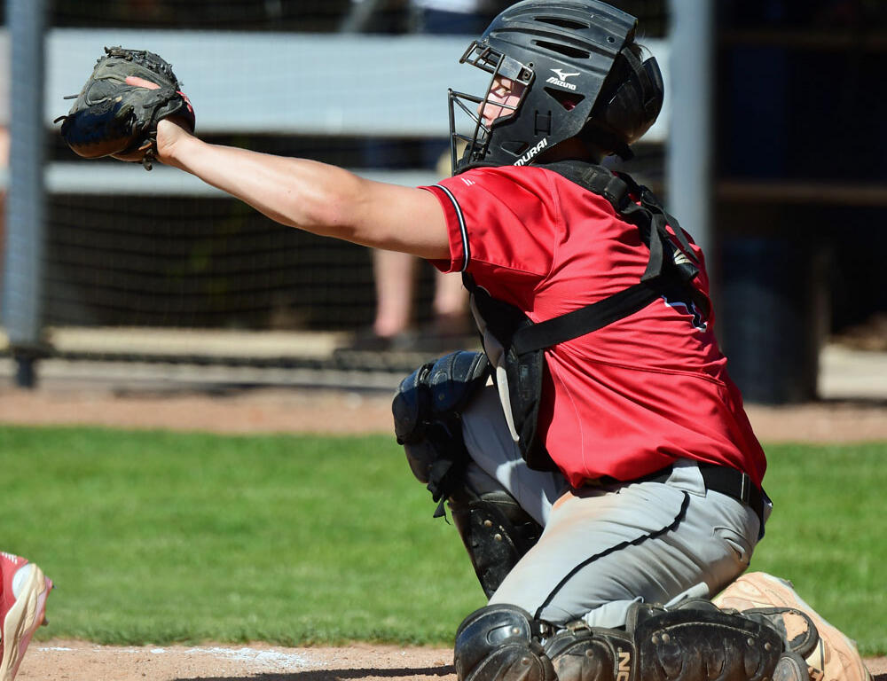 Baseball catcher in a red uniform kneels on dirt, glove extended to tag a runner at home plate.
