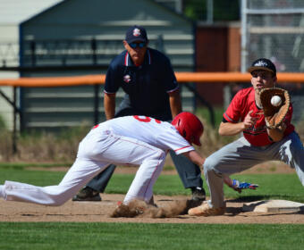 Baseball action: a runner slides into a base as a fielder in red aims to tag him, with an umpire watching in the background.