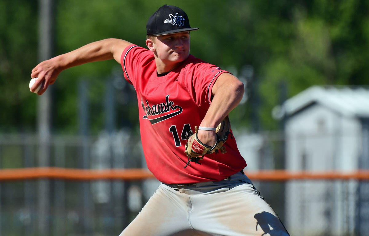 Baseball pitcher in a red jersey (number 14) and black cap delivering a pitch on a sunny field, with a fence and trees in the background.