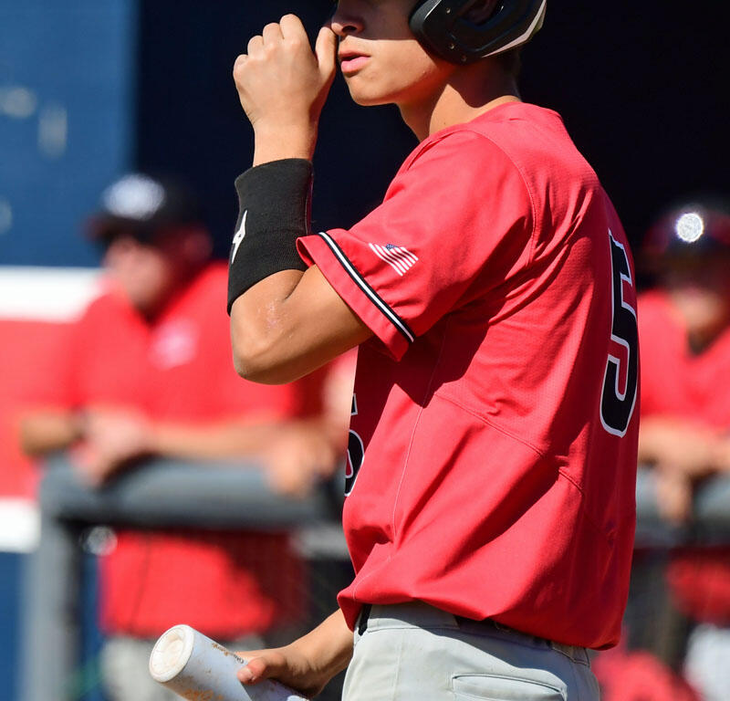 Baseball player in a red uniform and navy helmet touches his nose while gripping a bat, prepared at the dugout area.