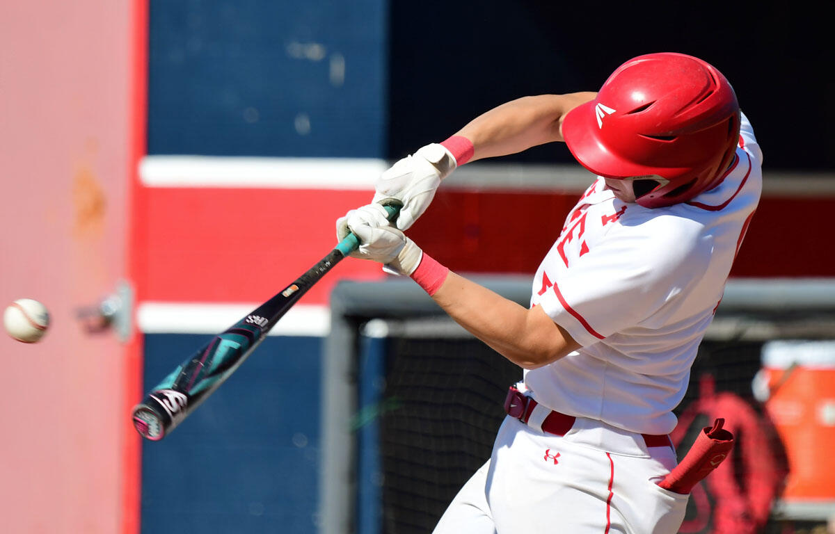Baseball batter in white and red swings a bat at a pitched ball with a red helmet on, action shot at the plate.