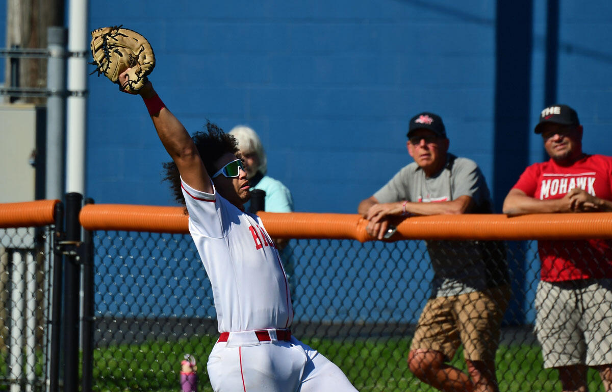 Baseball outfielder in white-red uniform leaps with glove raised to catch near the orange fence while spectators watch