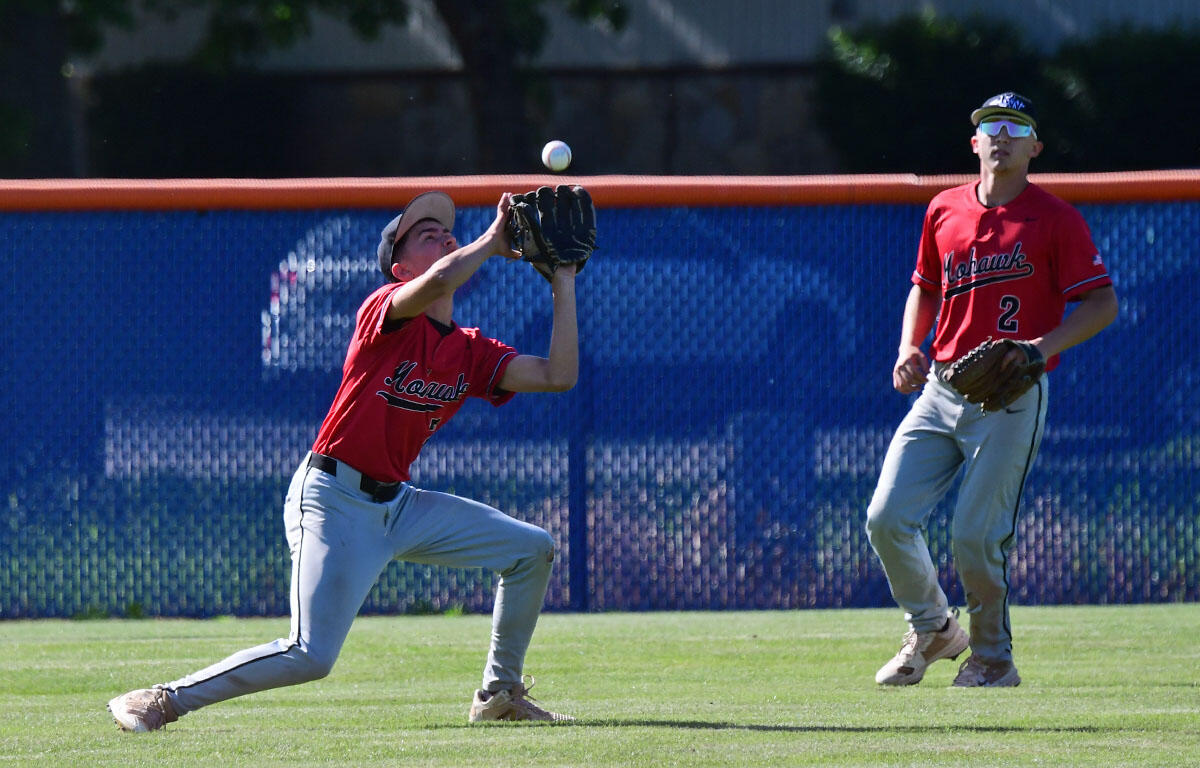 Baseball players in red jerseys on a grassy field; one fielder dives with glove up to catch a ball in midair while a teammate watches nearby.