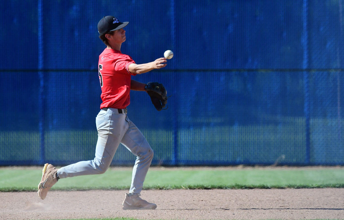 Baseball pitcher in a red jersey mid-pitch on the infield, blue outfield fence in the background.