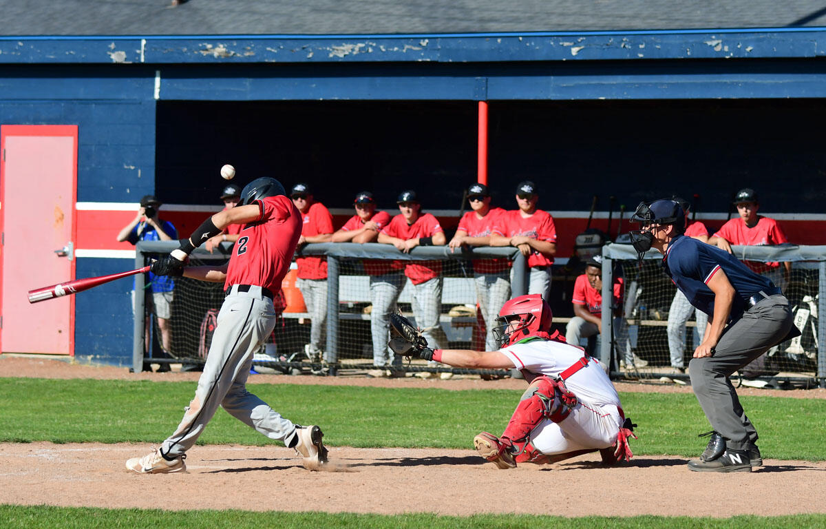 Baseball batter in red swings as the ball sails toward the plate, with catcher diving and reaching for the ball behind him.