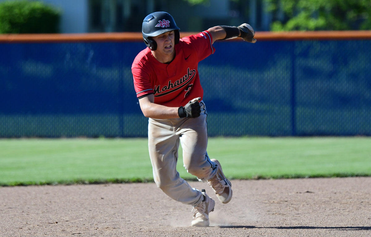 Baseball player in a red Mohawks jersey running on the infield dirt during a game, helmet on, focused on the play.