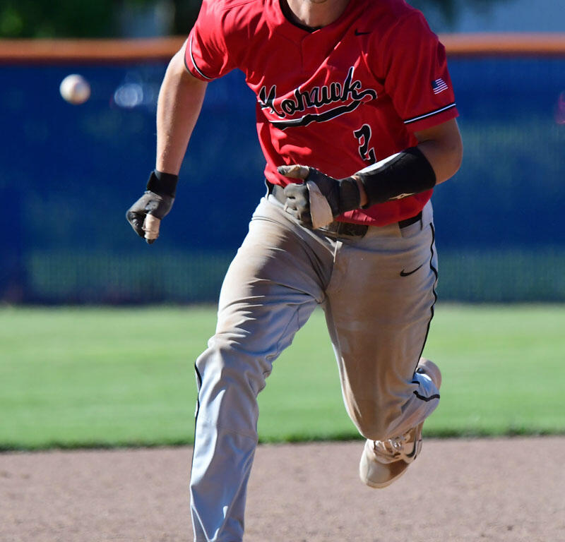 Baseball player in a red uniform and helmet sprinting on the infield as a ball bounces nearby on the dirt field.