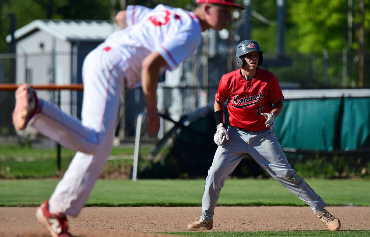 Two youth baseball players on a sunny field: a blur of a runner in white foreground while a red-jersey player with a helmet stands on the infield dirt, ready to field.