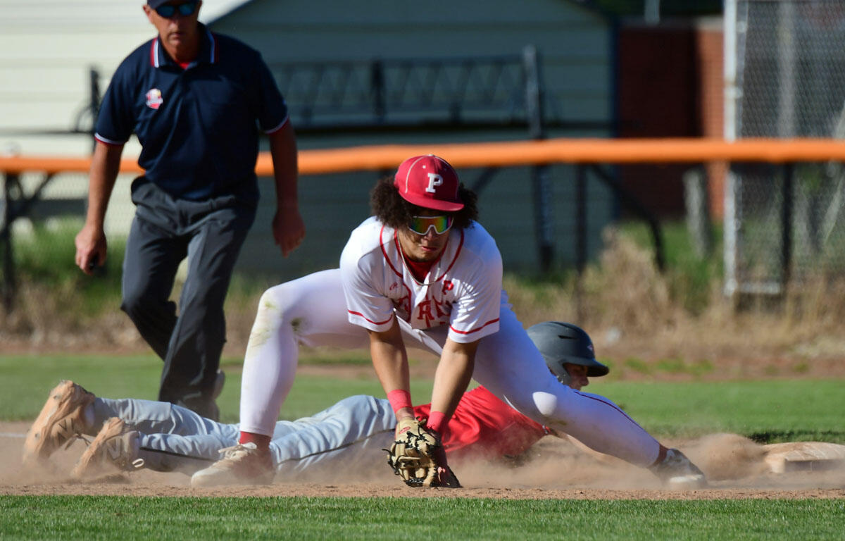 Baseball infielder in white and red, wearing a red cap and sunglasses, reaches for a ground ball as dust rises from a sliding baserunner nearby.