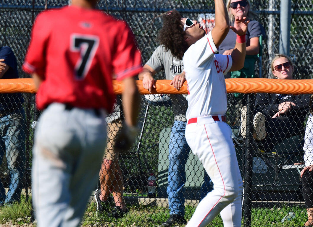 Baseball player in white and red jumps to catch a high popup near the chain-link fence as spectators watch nearby.