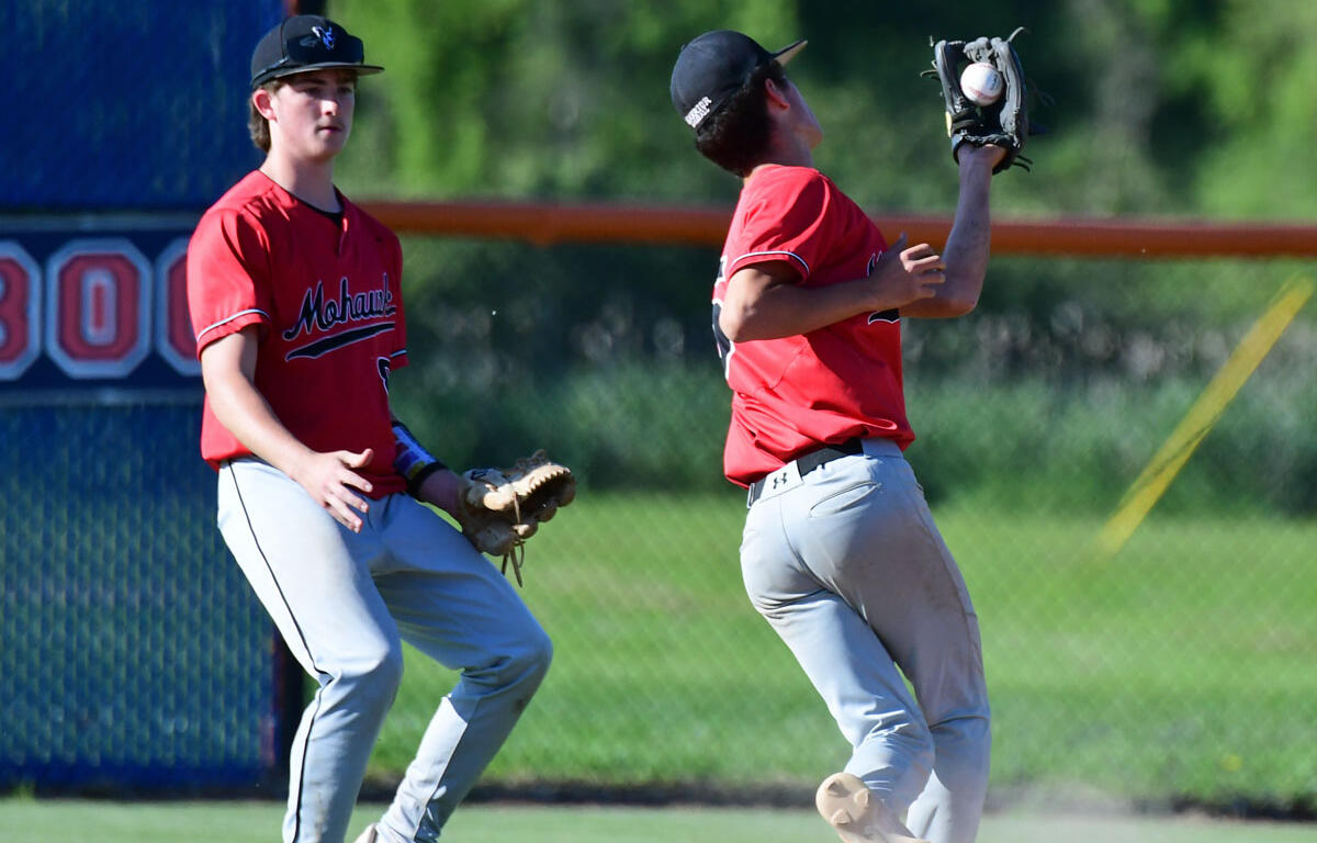 Two young baseball players in red uniforms on a sunny field; one reaches with his glove to catch a ball while the other watches nearby.