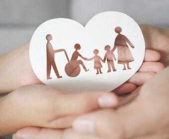 Paper heart cutout showing a family (parents, two children) holding hands, cradled in cupped hands to symbolize family support and care.