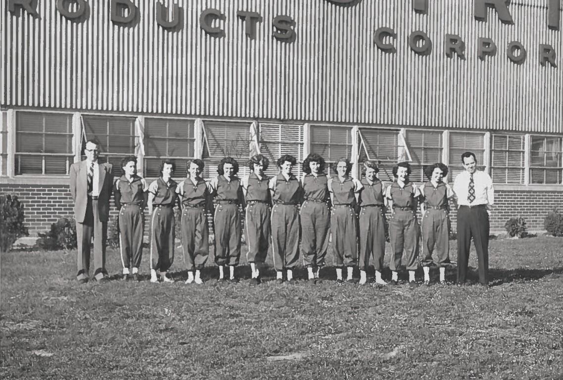A group of students in matching jumpsuits standing in a line with two men in business attire at each end, in front of a brick school building with a striped metal facade reading '... ROBOTICS ...' (black-and-white photo).