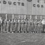 A group of students in matching jumpsuits standing in a line with two men in business attire at each end, in front of a brick school building with a striped metal facade reading '... ROBOTICS ...' (black-and-white photo).