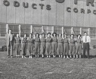 A group of students in matching jumpsuits standing in a line with two men in business attire at each end, in front of a brick school building with a striped metal facade reading '... ROBOTICS ...' (black-and-white photo).