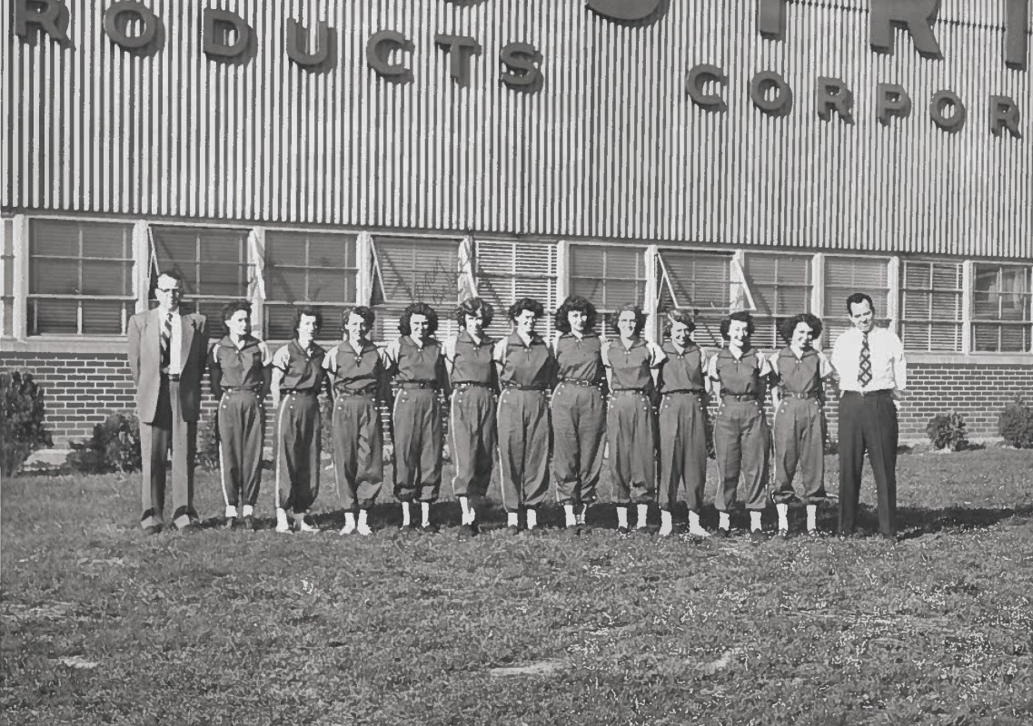 A group of students in matching jumpsuits standing in a line with two men in business attire at each end, in front of a brick school building with a striped metal facade reading '... ROBOTICS ...' (black-and-white photo).