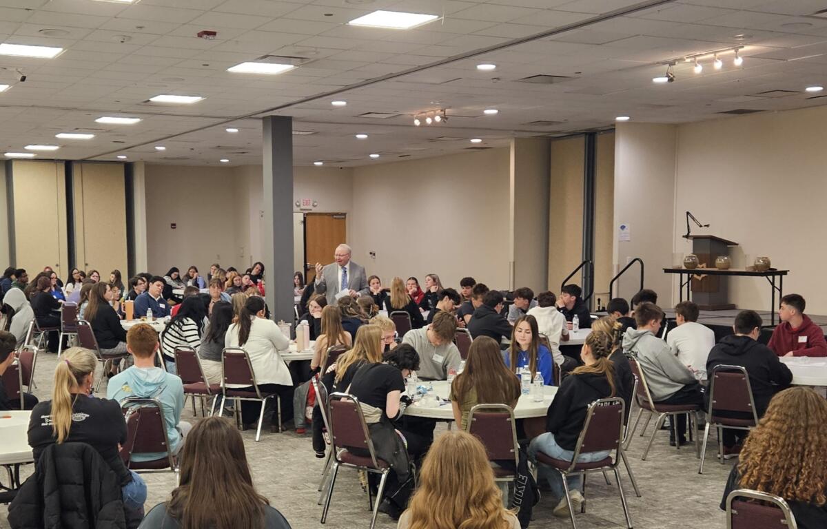 Male presenter in a suit addressing a large group of students seated at round tables in a conference room.