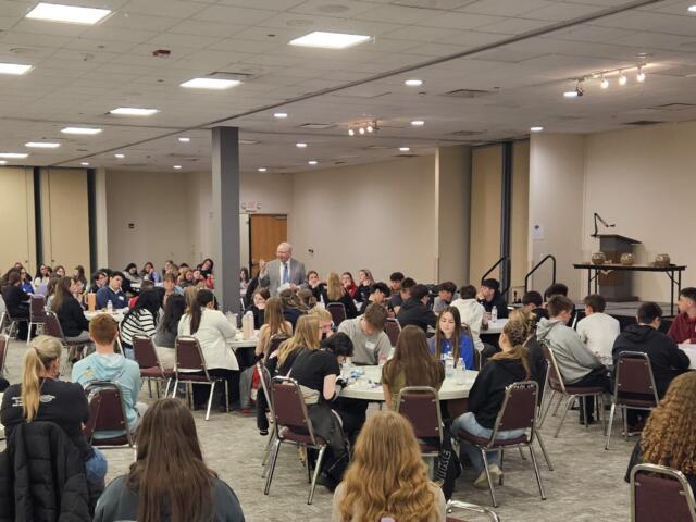 Male presenter in a suit addressing a large group of students seated at round tables in a conference room.