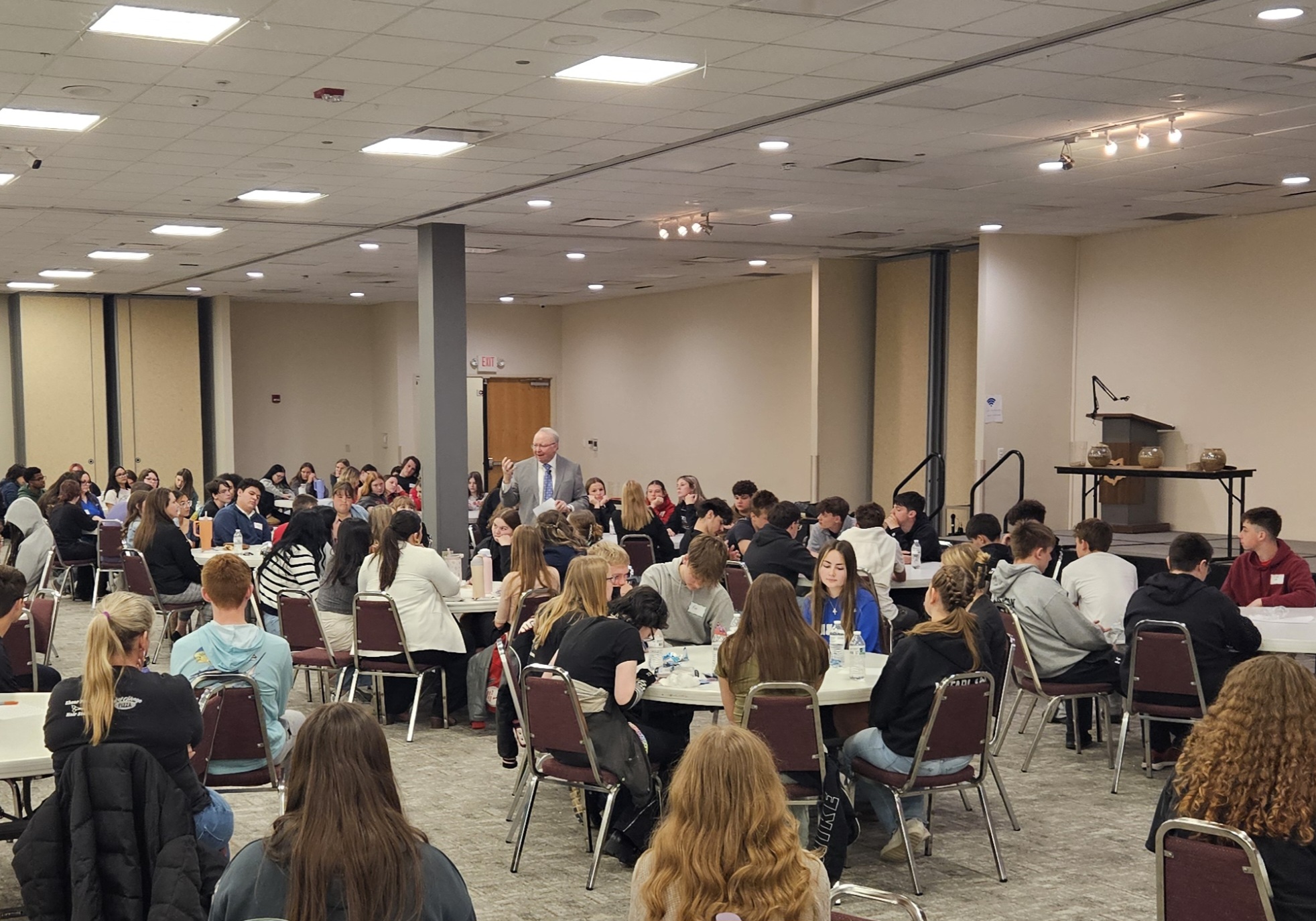 Male presenter in a suit addressing a large group of students seated at round tables in a conference room.