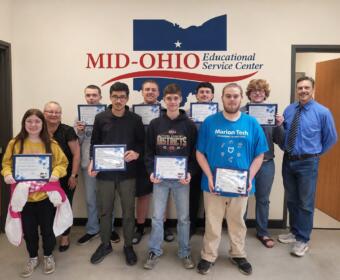 Group of students and two adults posing with certificates in a hallway at Mid-Ohio Educational Service Center.