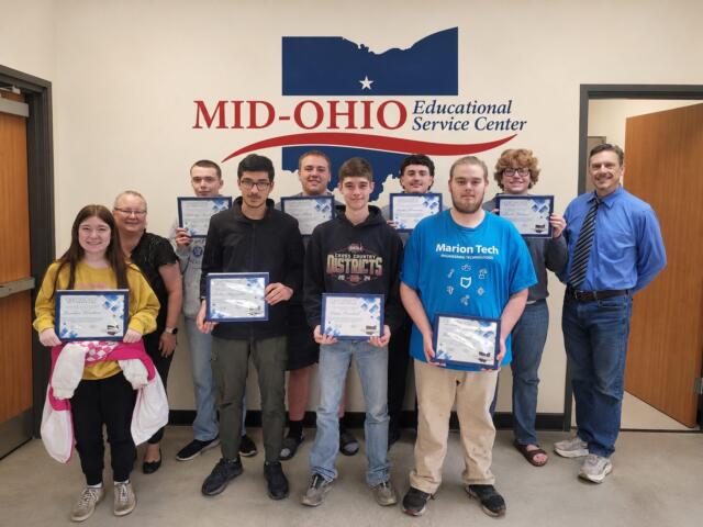 Group of students and two adults posing with certificates in a hallway at Mid-Ohio Educational Service Center.