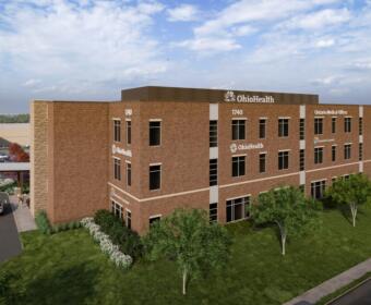 Three-story brick OhioHealth medical office building with parking lot and landscaped lawn, viewed from the grass.