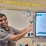 Smiling teacher in a classroom points to a small LED-lit microcontroller while a monitor shows Arduino code.