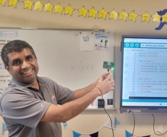 Smiling teacher in a classroom points to a small LED-lit microcontroller while a monitor shows Arduino code.