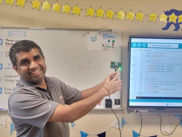 Smiling teacher in a classroom points to a small LED-lit microcontroller while a monitor shows Arduino code.