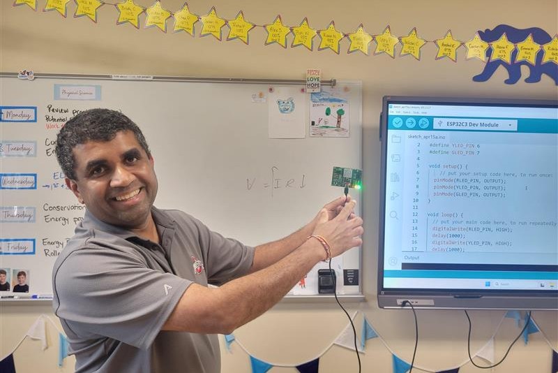 Smiling teacher in a classroom points to a small LED-lit microcontroller while a monitor shows Arduino code.