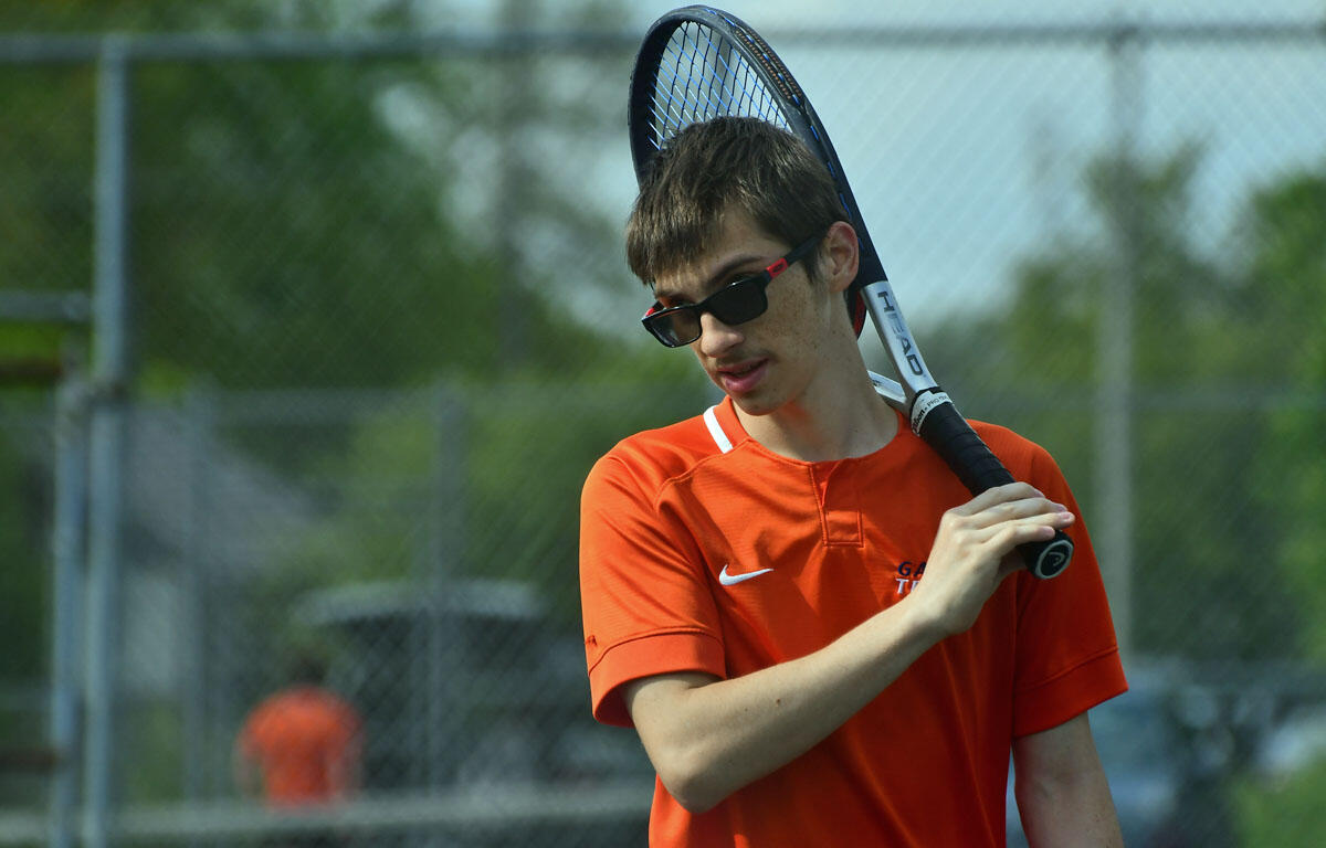 Teenage boy in an orange shirt and sunglasses holding a tennis racket on an outdoor court.