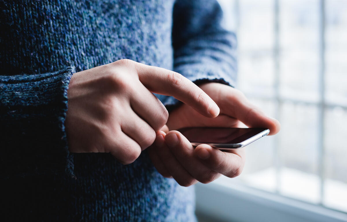 Person in a blue sweater using a smartphone, fingers tapping the screen near a window side light.