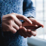 Person in a blue sweater using a smartphone, fingers tapping the screen near a window side light.