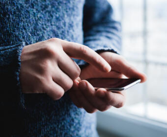 Person in a blue sweater using a smartphone, fingers tapping the screen near a window side light.