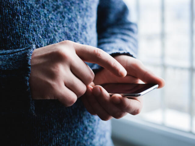 Person in a blue sweater using a smartphone, fingers tapping the screen near a window side light.
