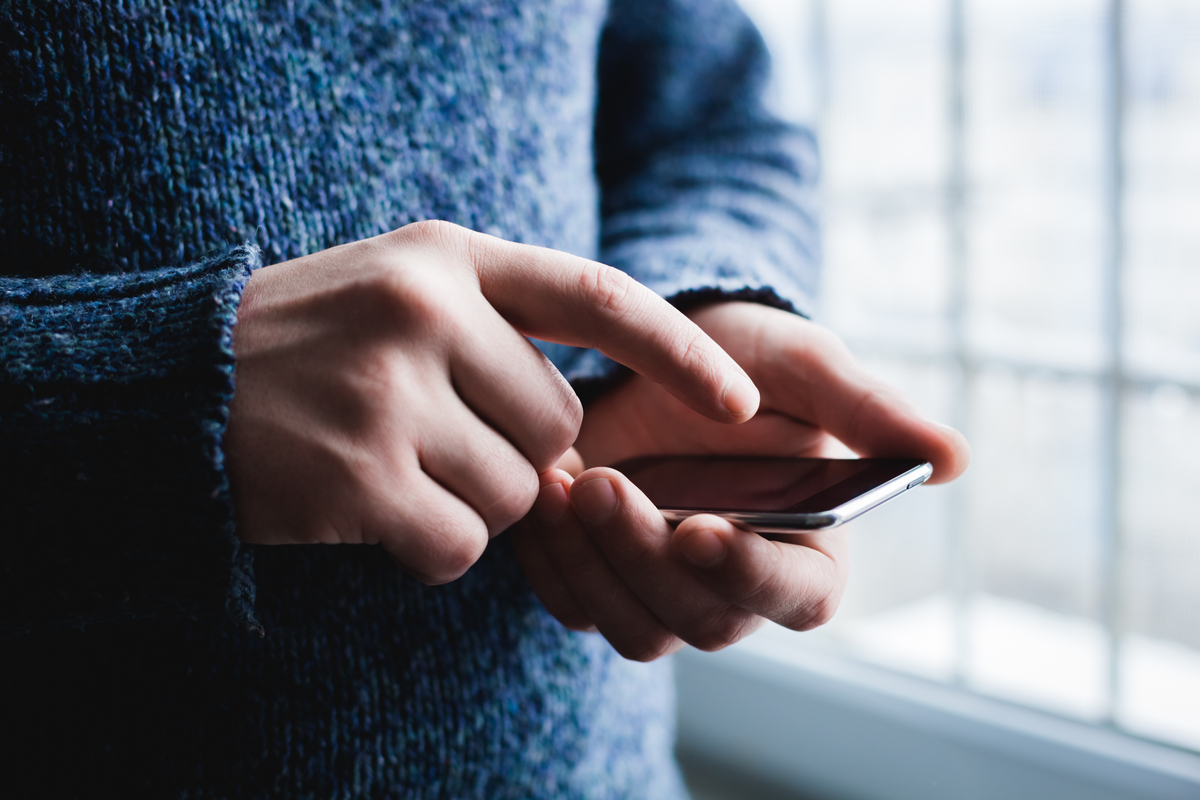Person in a blue sweater using a smartphone, fingers tapping the screen near a window side light.