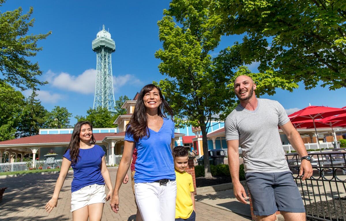 Family of four walking together outdoors on a sunny day near a tower, smiling and holding hands under green trees.