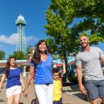 Family of four walking together outdoors on a sunny day near a tower, smiling and holding hands under green trees.