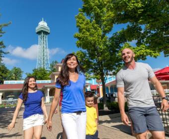 Family of four walking together outdoors on a sunny day near a tower, smiling and holding hands under green trees.