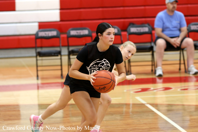 Girl in a black T-shirt dribbles a basketball during a youth game, red bleachers in the background.