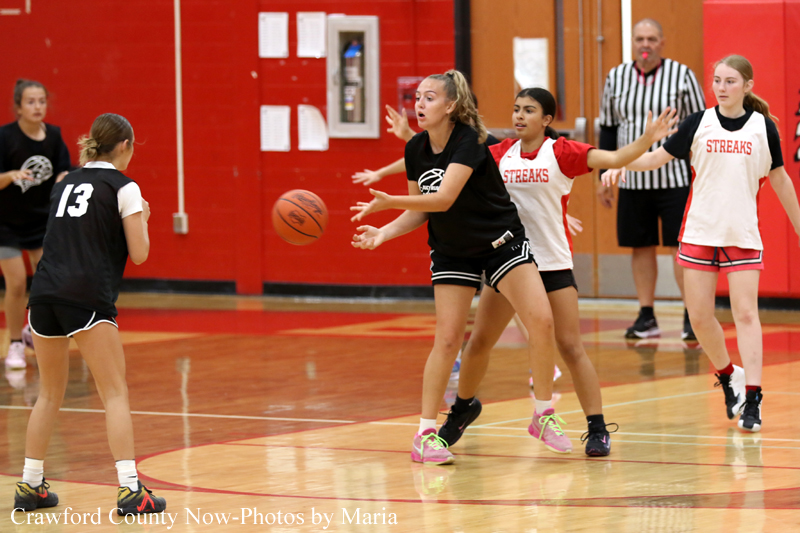 Girls' basketball game in a gym; player in black reaches for the ball while defenders in white 'STREAKS' jerseys converge nearby.