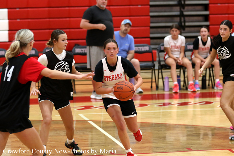 Girls' basketball game: central player in a white jersey labeled 'STREAKS' dribbles toward the basket while a defender reaches in black, with teammates and coaches on the bench in the background.