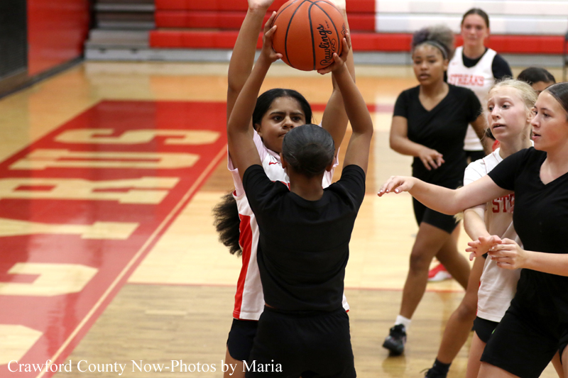 Girls basketball game in a gym; two players jump for the ball while teammates watch nearby.