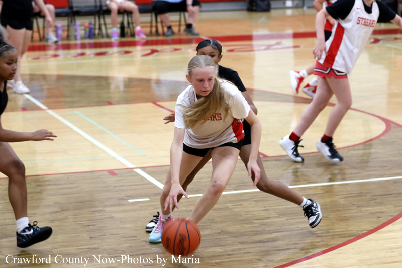 Girl in a white-and-red uniform dribbles a basketball with a defender close behind on an indoor court during a girls' basketball game in progress, other players in the background.