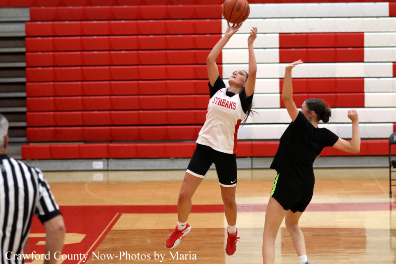 Two female players compete for a basketball in a gym; one in white jersey leaps for a shot while the other defends nearby. A referee is visible at left edge.