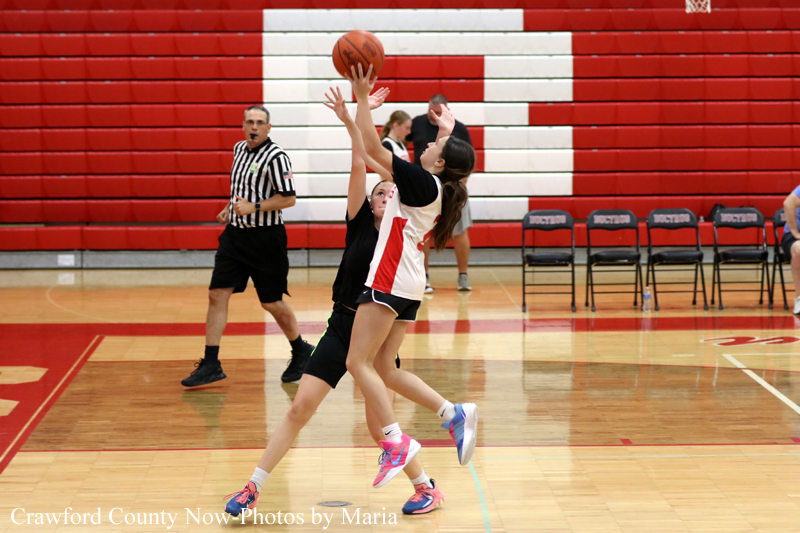Two girls jump for a basketball during a game in a gym with red and white bleachers; the referee watches nearby.,