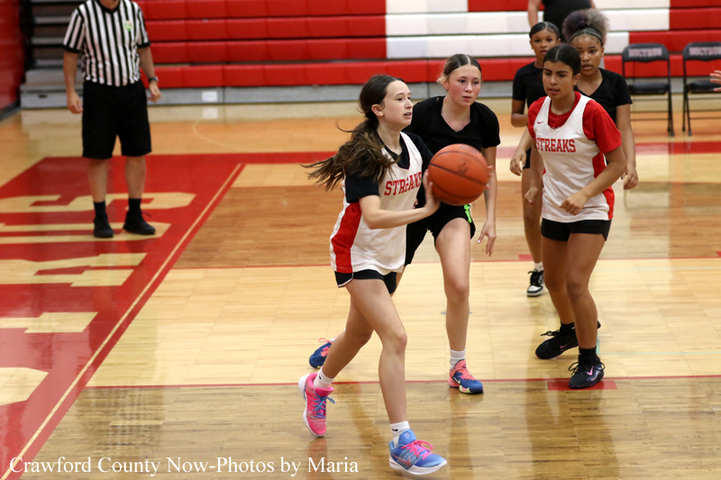 Girls' basketball game in a gym; a player in a white-red uniform dribbles the ball while teammates and defenders close in nearby.